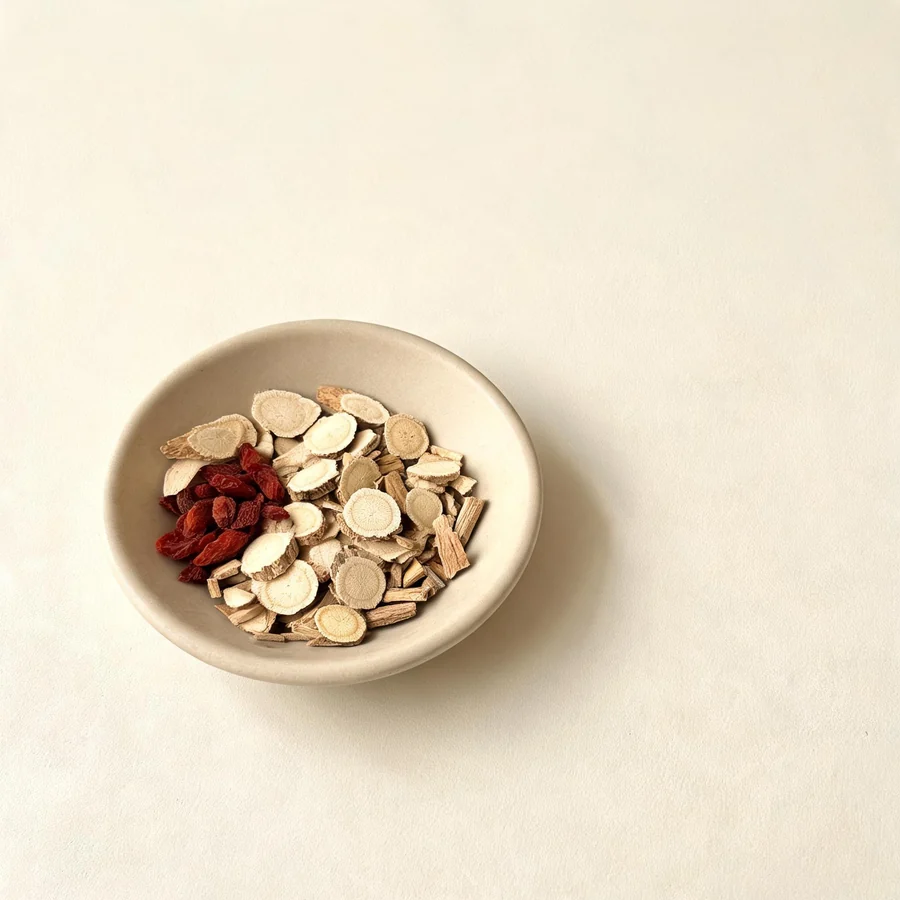 An editorial still life of the traditional Chinese herbs used for this condition, arranged on a warm off-white surface: pale root slices, a.